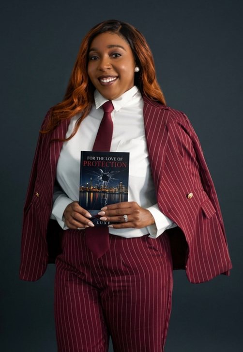 Luxe standing in a burgundy pinstripe suit, holding a copy of For The Love of Protection.